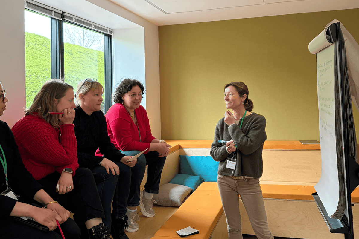 Une animatrice debout présente un paperboard à quatre femmes assises lors d'un atelier collaboratif dans une salle moderne - Fond'Actions des Possibles