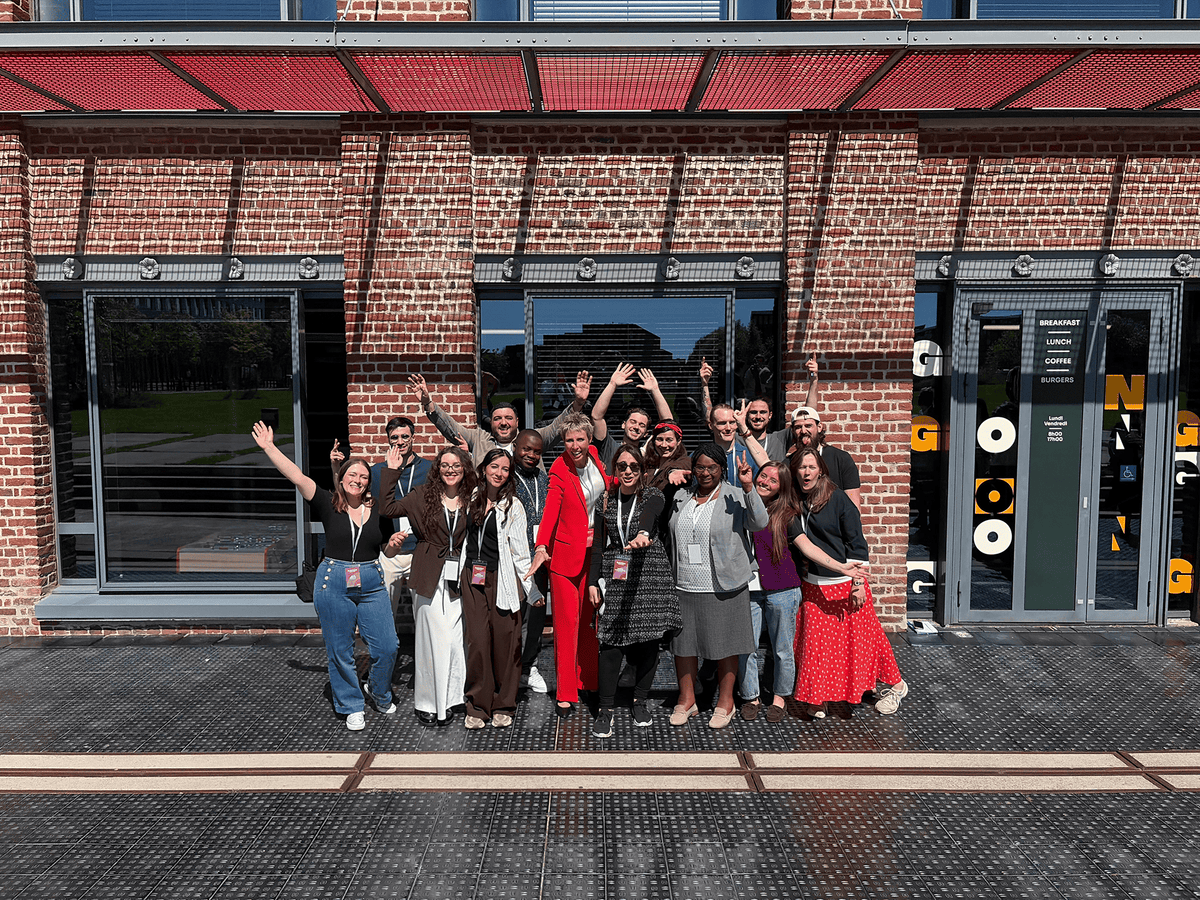 Une quinzaine de collaborateurs souriants et saluant de la main, posant pour une photo de groupe devant un bâtiment moderne en briques rouges - Fond'Actions des Possibles